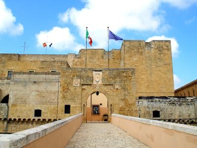 A stone fortress entrance with a bridge leading to an arched gateway, featuring Italian and EU flags atop the walls and a coat of arms above the arch.