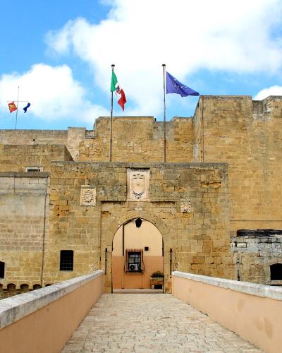 A stone fortress entrance with a bridge leading to an arched gateway, featuring Italian and EU flags atop the walls and a coat of arms above the arch.