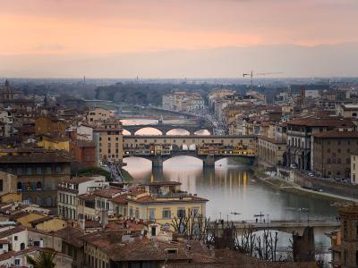 A high-angle view of the Ponte Vecchio bridge spanning the Arno River in Florence, Italy, with the cityscape and distant hills under a sunset sky.