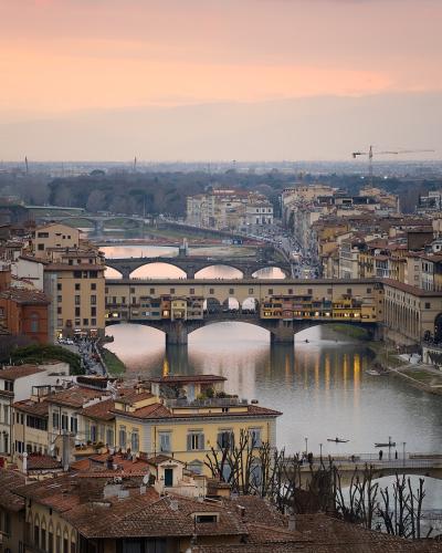 A high-angle view of the Ponte Vecchio bridge spanning the Arno River in Florence, Italy, with the cityscape and distant hills under a sunset sky.