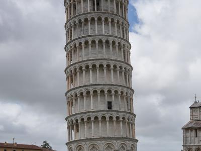 A low-angle photograph of the Leaning Tower of Pisa, a white marble Romanesque bell tower with eight stories of arcades, leaning noticeably to the right, topped with a red flag, surrounded by tourists on a green lawn under a cloudy sky, with part