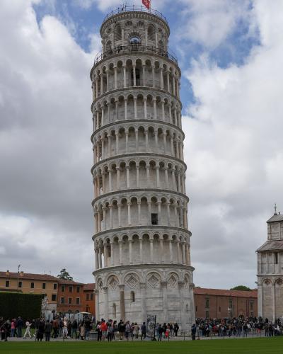 A low-angle photograph of the Leaning Tower of Pisa, a white marble Romanesque bell tower with eight stories of arcades, leaning noticeably to the right, topped with a red flag, surrounded by tourists on a green lawn under a cloudy sky, with part