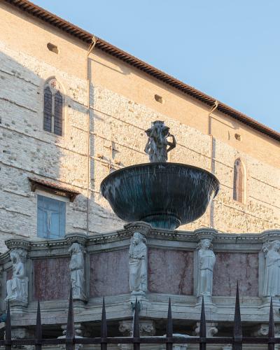 A large, ornate medieval stone fountain with a central bronze basin and statue, surrounded by sculpted figures on a hexagonal base, situated in front of a historic stone building with arched windows under a clear sky.