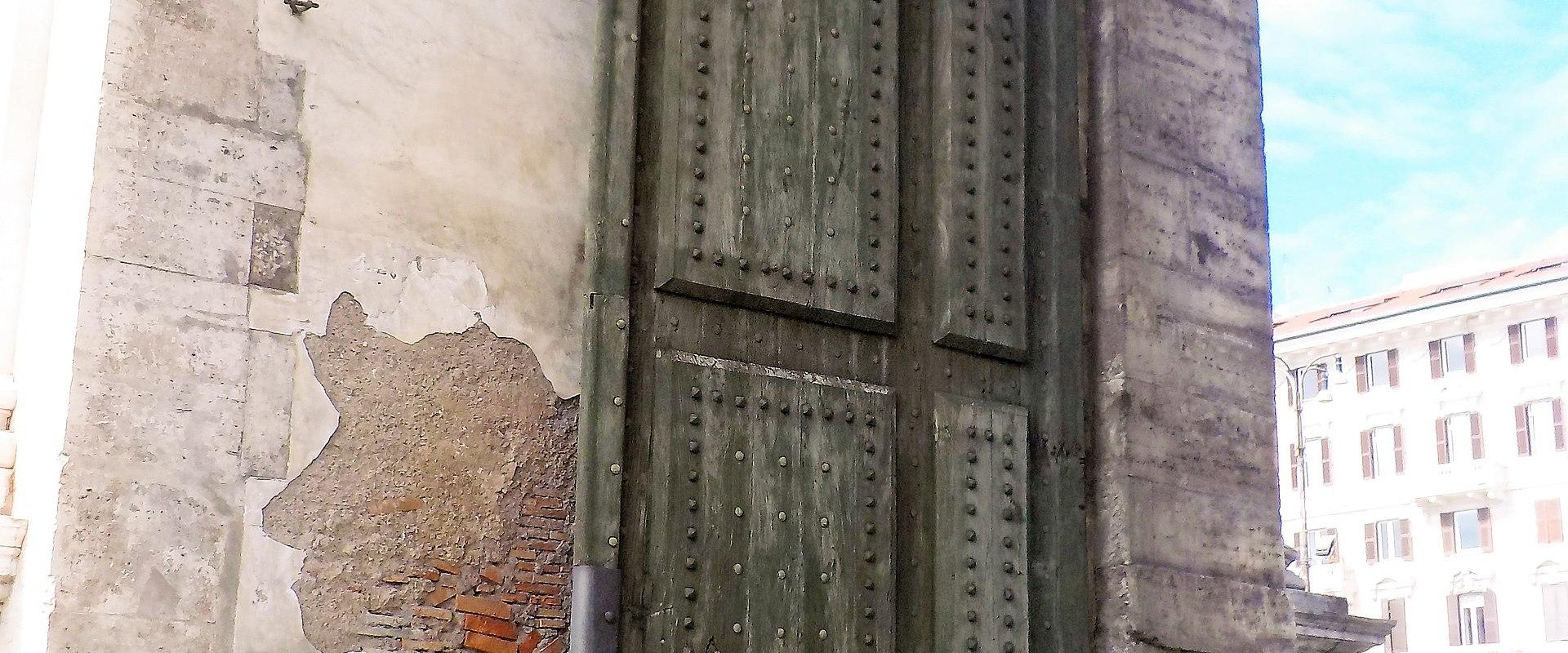 A close-up view of a massive, studded wooden door set within a stone archway, with a smaller pedestrian door to the left bearing graffiti and people visible in the background on a cobblestone street.