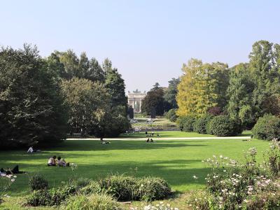 A wide view of a large, manicured green park with people relaxing on the grass, framed by trees, with the Arco della Pace triumphal arch visible in the distance.