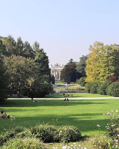 A wide view of a large, manicured green park with people relaxing on the grass, framed by trees, with the Arco della Pace triumphal arch visible in the distance.