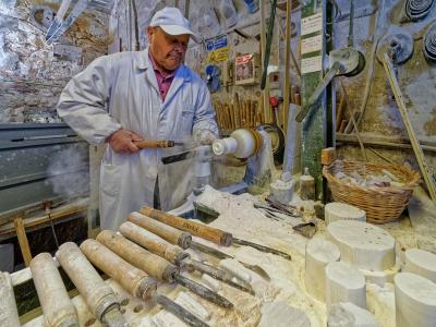 A craftsman in a white coat and cap operates a lathe to shape a piece of white alabaster in a workshop filled with tools, raw stone blocks, and dust; signage on the wall includes "LA DOLCE..." and safety warnings.