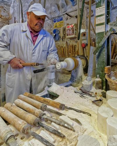 A craftsman in a white coat and cap operates a lathe to shape a piece of white alabaster in a workshop filled with tools, raw stone blocks, and dust; signage on the wall includes "LA DOLCE..." and safety warnings.