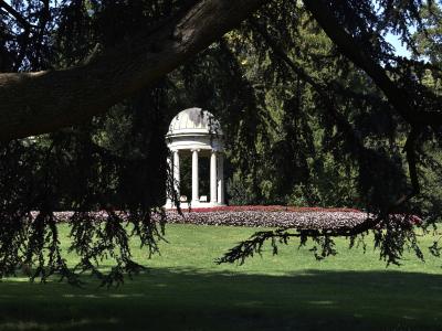 A white neoclassical temple-like structure (tempietto) with columns and a dome, situated in a manicured park with flowerbeds and framed by tree branches.
