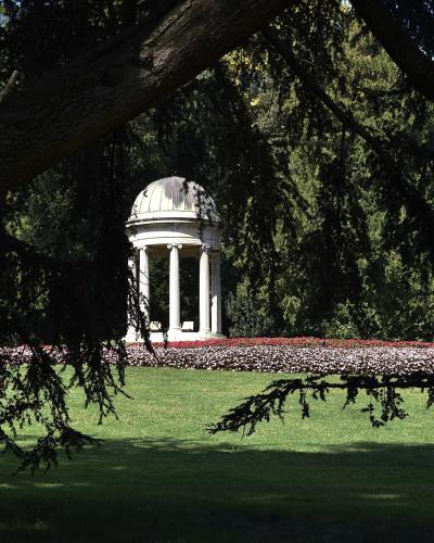 A white neoclassical temple-like structure (tempietto) with columns and a dome, situated in a manicured park with flowerbeds and framed by tree branches.