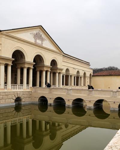 A Renaissance palace featuring a long loggia with arches and columns, situated next to a rectangular reflecting pool with arched openings in its retaining wall.
