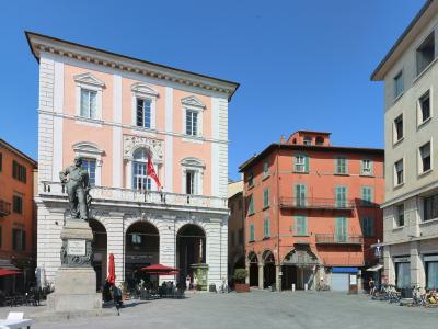 The image shows a sunlit Italian piazza featuring a prominent pink and white building with arched ground-floor loggias, a statue of a man on a pedestal in the foreground, and surrounding multi-story buildings with shuttered windows under a clear b