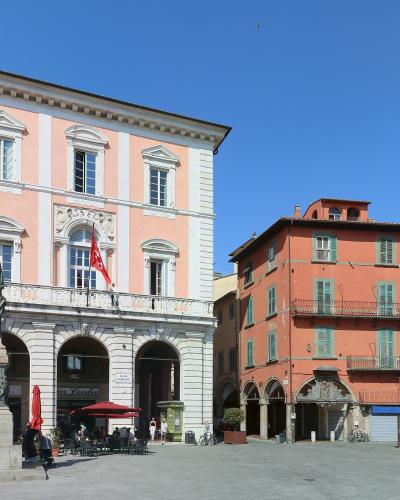 The image shows a sunlit Italian piazza featuring a prominent pink and white building with arched ground-floor loggias, a statue of a man on a pedestal in the foreground, and surrounding multi-story buildings with shuttered windows under a clear b