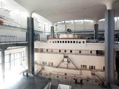 The image shows the interior of a large museum hall featuring a massive white ship hull as the central exhibit, with Italian text on a banner reading "...tà che schiude la fantasia..." and industrial-style architecture with high ceilings and suppo