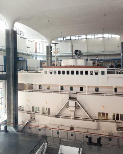 The image shows the interior of a large museum hall featuring a massive white ship hull as the central exhibit, with Italian text on a banner reading "...tà che schiude la fantasia..." and industrial-style architecture with high ceilings and suppo
