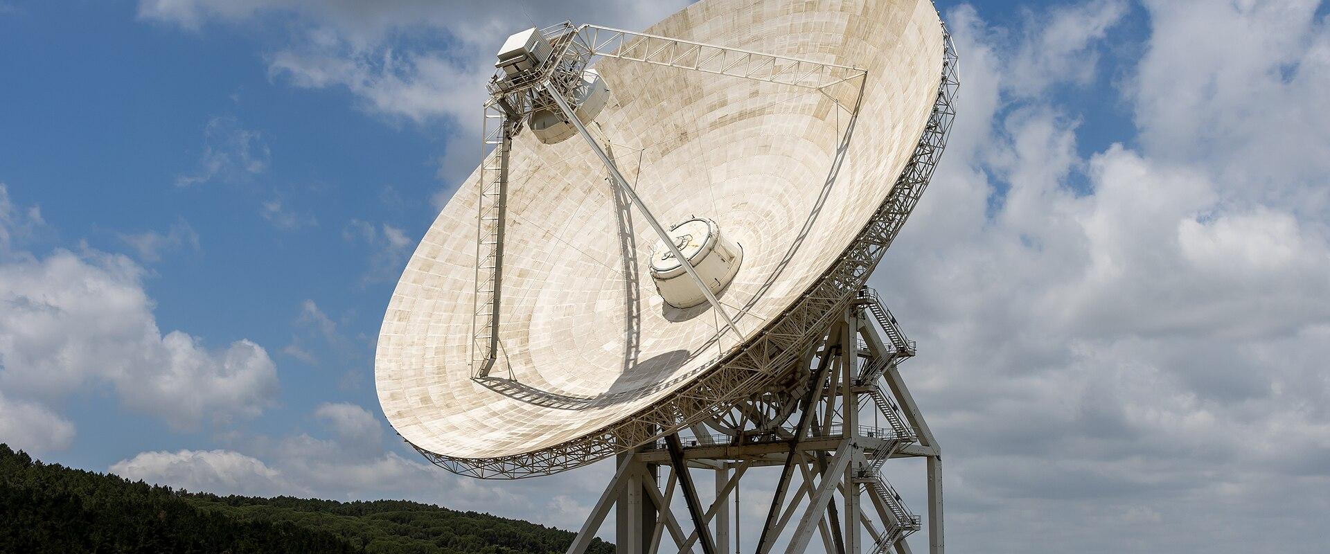 A large white radio telescope dish on a metal support structure in a grassy field with forested hills under a blue sky with clouds.