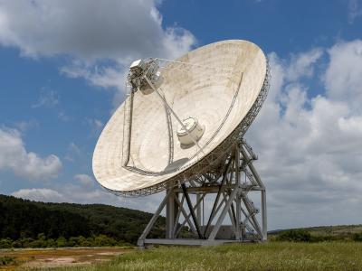 A large white radio telescope dish on a metal support structure in a grassy field with forested hills under a blue sky with clouds.