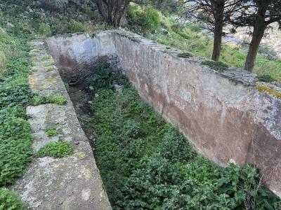 A weathered stone retaining wall or foundation of an old structure, overgrown with green vegetation, situated on a hillside with trees and a distant view of a town below.