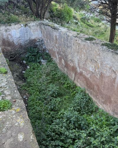 A weathered stone retaining wall or foundation of an old structure, overgrown with green vegetation, situated on a hillside with trees and a distant view of a town below.