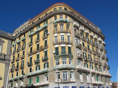 A large, multi-story yellow and grey building with a distinctive curved corner facade, green shutters, and numerous balconies, situated on a street corner under a clear blue sky.
