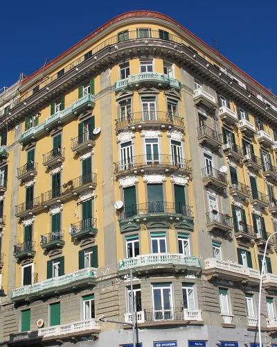 A large, multi-story yellow and grey building with a distinctive curved corner facade, green shutters, and numerous balconies, situated on a street corner under a clear blue sky.