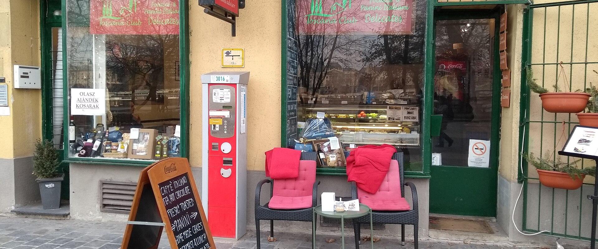 The image shows the exterior of a café or small restaurant named "Toscana Club," with signage in Hungarian (e.g., "OLASZ AJÁNDÉK KÜSZÖBÖK" meaning "Italian Gift Threshold," and menu items like "CAFFE ITALIANO," "PANINI," "GELATO") displayed on win