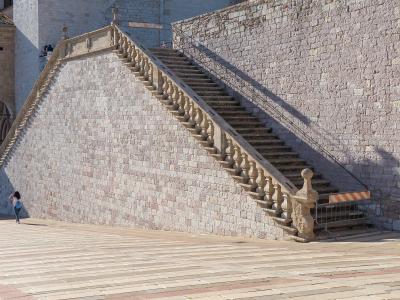 A long, steep outdoor marble staircase with a stone balustrade ascending a large stone retaining wall, with a paved plaza in the foreground and part of a Gothic-style building entrance visible on the left.