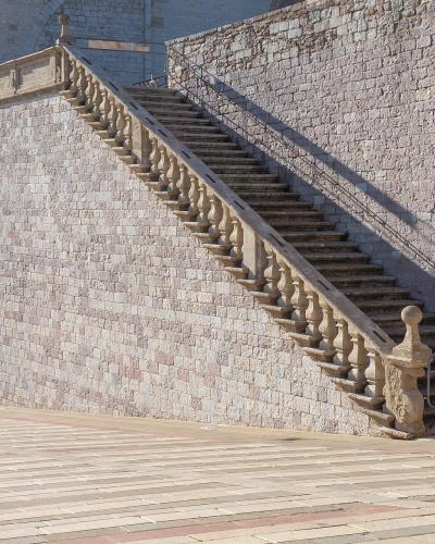 A long, steep outdoor marble staircase with a stone balustrade ascending a large stone retaining wall, with a paved plaza in the foreground and part of a Gothic-style building entrance visible on the left.