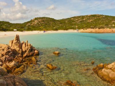 A scenic coastal view of a crescent-shaped white sand beach with turquoise water, framed by distinctive reddish-brown granite rock formations in the foreground and green-covered hills in the background; a few beachgoers and umbrellas are visible o