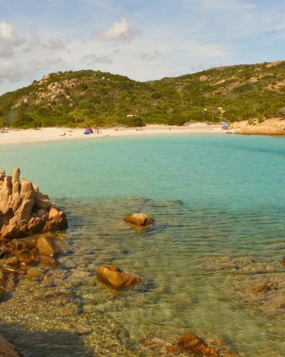 A scenic coastal view of a crescent-shaped white sand beach with turquoise water, framed by distinctive reddish-brown granite rock formations in the foreground and green-covered hills in the background; a few beachgoers and umbrellas are visible o