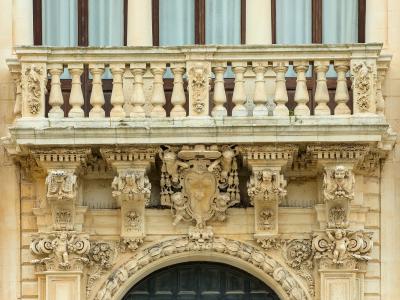 A close-up architectural detail of an ornate stone balcony and arched doorway featuring Baroque-style carvings, including cherubs, a central coat of arms, and balustrades on light-colored limestone.