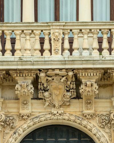 A close-up architectural detail of an ornate stone balcony and arched doorway featuring Baroque-style carvings, including cherubs, a central coat of arms, and balustrades on light-colored limestone.