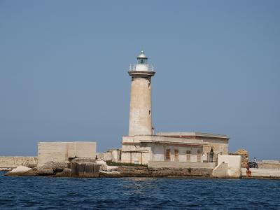 A tall, cylindrical lighthouse with a lantern room and gallery, situated on a stone breakwater or pier extending into blue water under a clear sky; no visible signage or text is present in the image.