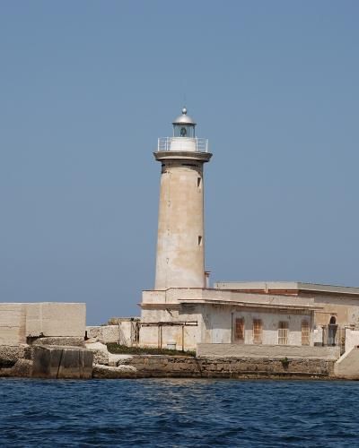 A tall, cylindrical lighthouse with a lantern room and gallery, situated on a stone breakwater or pier extending into blue water under a clear sky; no visible signage or text is present in the image.