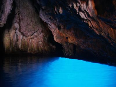 The image shows the interior of a sea cave with dark, textured limestone walls and a body of water glowing with an intense, unnatural electric blue light, likely from artificial underwater lighting; no signage or identifying features are visible.