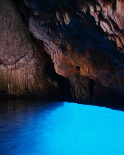The image shows the interior of a sea cave with dark, textured limestone walls and a body of water glowing with an intense, unnatural electric blue light, likely from artificial underwater lighting; no signage or identifying features are visible.