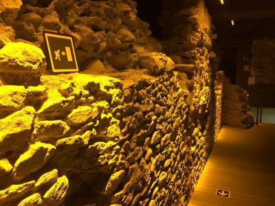 The image shows an interior view of a museum exhibit featuring exposed ancient stone walls, likely Roman ruins, illuminated by warm yellow lighting, with modern wooden flooring and directional signage (including an emergency exit sign and floor ar