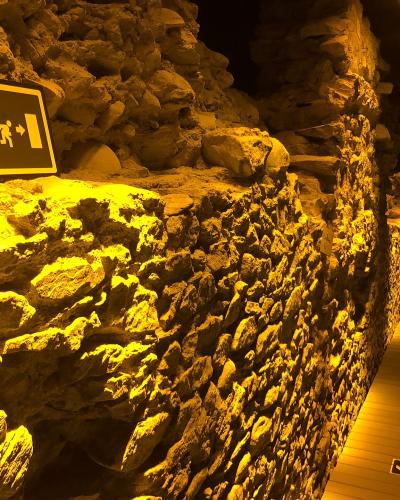 The image shows an interior view of a museum exhibit featuring exposed ancient stone walls, likely Roman ruins, illuminated by warm yellow lighting, with modern wooden flooring and directional signage (including an emergency exit sign and floor ar