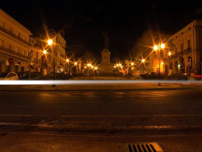 A night-time long-exposure photograph of a paved town square or piazza featuring a central monument on a pedestal, flanked by multi-story buildings and illuminated by streetlights that create starburst effects, with light trails from moving vehicl