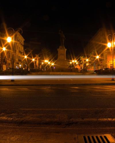 A night-time long-exposure photograph of a paved town square or piazza featuring a central monument on a pedestal, flanked by multi-story buildings and illuminated by streetlights that create starburst effects, with light trails from moving vehicl