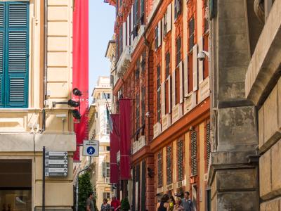 The image shows a narrow, cobblestone pedestrian street in an Italian city, flanked by tall, colorful historic buildings with shutters, featuring red banners that read "Palazzo Bianco" and a sign for "area pedonale" (pedestrian area).