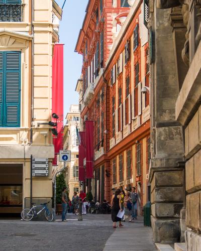 The image shows a narrow, cobblestone pedestrian street in an Italian city, flanked by tall, colorful historic buildings with shutters, featuring red banners that read "Palazzo Bianco" and a sign for "area pedonale" (pedestrian area).