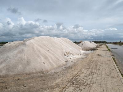 The image shows large conical mounds of white harvested salt situated next to a paved service road and shallow evaporation ponds under a cloudy sky.