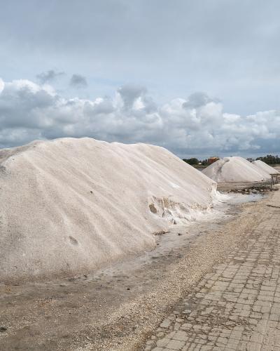 The image shows large conical mounds of white harvested salt situated next to a paved service road and shallow evaporation ponds under a cloudy sky.