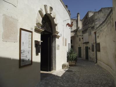 The image shows the entrance to a white-washed stone building with a distinctive arched doorway featuring carved stone details, a framed information board on the wall, and a narrow cobblestone alleyway; a sign hanging from a wire displays a rooste