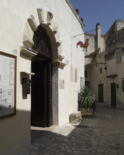 The image shows the entrance to a white-washed stone building with a distinctive arched doorway featuring carved stone details, a framed information board on the wall, and a narrow cobblestone alleyway; a sign hanging from a wire displays a rooste