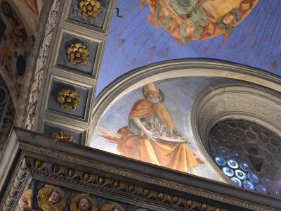 The image shows an interior ceiling fresco of a bishop holding a model city, adjacent to a decorative arch and a circular stained-glass window with blue glass discs.