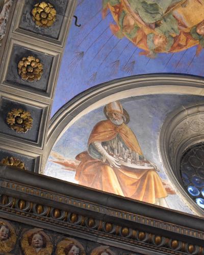 The image shows an interior ceiling fresco of a bishop holding a model city, adjacent to a decorative arch and a circular stained-glass window with blue glass discs.