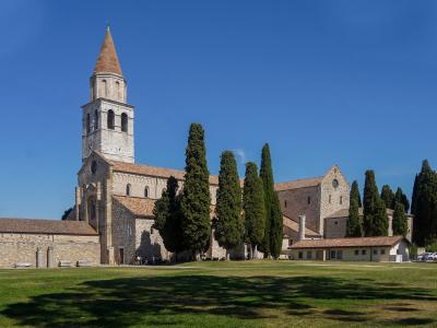 A large medieval stone basilica with a tall, conical-roofed bell tower and a row of cypress trees in front of a grassy field under a blue sky.