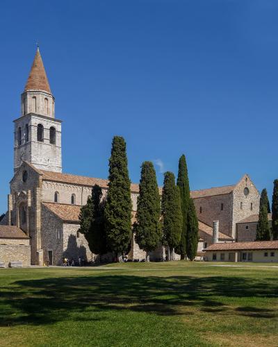 A large medieval stone basilica with a tall, conical-roofed bell tower and a row of cypress trees in front of a grassy field under a blue sky.